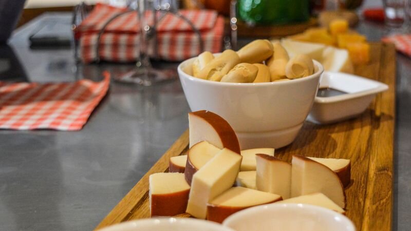 A wooden board with sliced cheese and a bowl of bread rolls, set alongside wine glasses