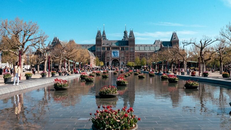 An ornate brick building with twin spired towers behind a reflecting pool lined with pots of red tulips
