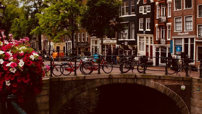 Bicycles parked along a canal bridge decorated with hanging flowers, with Amsterdam canal houses beyond